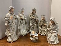 Six-piece nativity figurine set including baby Jesus in the manger and three wise men, Mary, and Joseph, all painted in white and silver with decorative details, displayed on wooden surface.