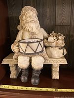 Front view of Santa Claus figurine seated on a bench holding a drum with toy sack beside him.