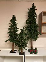 Full view of three artificial Christmas trees placed on white shelving against a beige wall, showing varying sizes and two small decorative bundles at base.