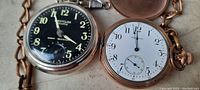 Two old pocket watches side by side showing their faces; one black dial Westclox Scotty and one white dial goldtone watch with loose etched cover.