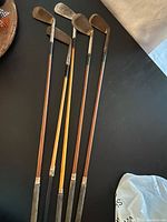 Full view of 5 antique golf clubs with wooden shafts laid on dark surface showing entire club lengths