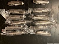 Twelve stainless steel pocket knives with corkscrews in individual plastic packaging, displayed on a dark surface.