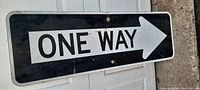 Photo shows older metal street sign with black background and white text reading 'ONE WAY' with arrow pointing right.