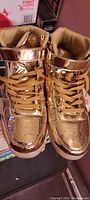 Pair of gold metallic high top tennis shoes shown from an oblique front angle. Shoes have laces and velcro strap, shiny reflective gold finish, with lightly used soles.