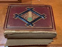 Cover of antique book with maroon leather, embossed geometric and floral design in gold and blue, showing heavy wear and damage on spine.