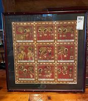 Full front view of framed artwork with nine panels and inscriptions. Shows entire piece with light reflections visible on glass.