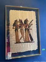 Framed artwork showing three Egyptian figures on papyrus with metal frame and glass.