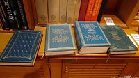 Four hardcover books with ornate blue and green covers displayed on a wooden shelf.