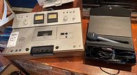 Front view of Akai cassette deck with control panel, VU meters, and cassette compartment along with Monacor microphone and case on wooden surface.