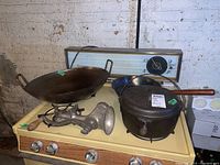 Photo showing the cast iron wok on left, metal universal grinder in front, cast iron pot with lid and chestnut roasting pan with wooden handle, all placed on an old stove.