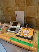 Photo showing open Nintendo DS Lite console, Brain Age game case, instruction booklet, and power adapter on tiled countertop.