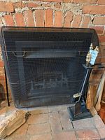 Set of black metal fireplace tools on stand with brass finials, a hand broom, and a mesh fireplace screen in front of a brick fireplace.