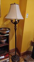Full view of the floor lamp against a yellow wall showing the base, pole, and fabric shade with braided trim.