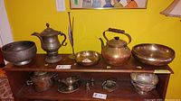 Overview of the copper kettle, metal coffee pot, three metal bowls of varying sizes, and smaller metal serving pieces arranged on wooden shelf.