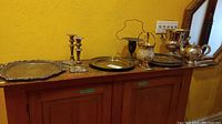 Wide view of various silver plated items laid out on wooden surface against yellow wall. Shows multiple trays, ice buckets, teapot vase, and candlesticks.