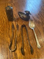 Full view of the lot showing the two pairs of repeating silver tongs, silver spoon with ornate handle, 90% silver duck vessel, and cylindrical jar with floral embossing.