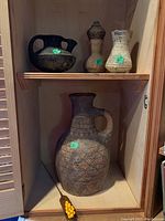 Three clay vases and one jug displayed inside a wooden cabinet shelf. Large vase is 16 inches tall with handle and textured pattern. Smaller clay jug and vases are above and beside it. Cluster of yellow-orange decorative glass berries at bottom.