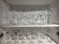 Wide view of two shelves containing a total of 19 clear glass wine and liqueur glasses of various styles and sizes arranged in rows.