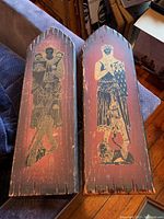Two wooden plaques with pointed arch tops, each featuring a brass rubbing of a medieval figure with shield, on a reddish-brown wood background.
