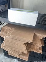 Flat packed brown corrugated cardboard shipping boxes stacked on a metal shelf with one white assembled box on top for size comparison.