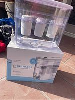 Photo of the clear plastic alkaline water dispenser with white lid and three visible filter cartridges inside. Unit is placed on top of its product box showing branding and product details.