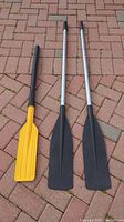 Three paddles laid on brick pavement: two aluminum paddles with black handles and blades, and one yellow plastic paddle with black handle