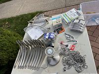 Nine silver shelf brackets, various styles including five IKEA Ekby shelf brackets, high pot light fixture, hooks, chains and other small hardware on table outside.