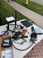 Photo displaying assorted camera gear on a white table including camera tripod with LED light panel, vintage Keystone XR camera, various cables, microphone, phone holder, tablet case, and DVD writer device.