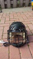 Front view of black Itech hockey helmet with metal face cage and chin strap on brick pavement