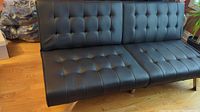 Front angled view of the black tufted futon couch showing seat and back cushions with chrome legs on wood floor.