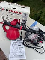 Complete lot showing the Scunci branded box, red and black steam cleaner unit with accessories and instruction manual outside the box.