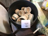 Top view of black 5-gallon bucket filled with new beige ABS plumbing fittings, including elbows and couplings, placed on a chair.