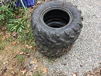 Two used ATV tires stacked vertically on gravel with surrounding greenery, showing the tread and size markings.