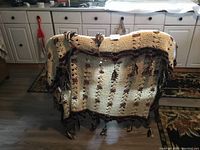 Large crocheted blanket draped over chair showing cream and brown color with tassels on edges.