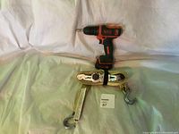 Black & Decker cordless drill and coiled beige ratchet strap with metal hooks seen from above on a white fabric background.