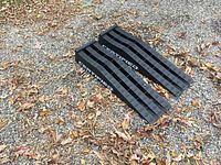 Pair of black plastic car ramps photographed from above on gravel with fallen leaves. 'CERTIFIED' branding visible on sides.
