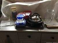 Full view of the lot showing stainless steel kettle, metal grater, food storage containers, springform pan, pasta boat, black baking trays, and clear glass pitcher