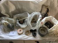Plastic bags holding brass and metal plumbing fittings, rolls of plumber's tape, pipe clamps, and other plumbing parts arranged on a surface.
