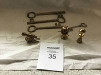 Five brass skeleton keys alongside three brass miniatures: a cannon, an oil lamp, and a candlestick, placed on a light cloth.