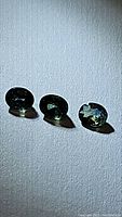 Three oval cut multicolored blue-green natural sapphires arranged in a row on a white textured background, showing their side profile and brilliance under light.