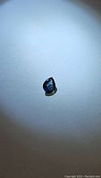 Photo of single pear cut blue green sapphire gemstone on white surface under light, showing color and cut details.