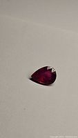 Photo of a deep red pear shaped ruby gemstone on plain light surface, shows rich color and shape clearly.