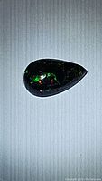 Pear-shaped jet black Ethiopian opal with green and red color flashes on a light background, showing the top view of the cabochon.