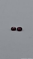 Pair of two deep pink to electric purple oval garnet gemstones on a gray background, showing color and size details.