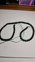 Full strand of dark green spherical emerald beads on a white background, showing the entire length of the strand and the green thread stringing the beads.