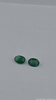 Two oval cut green gemstones placed on a flat white surface, showing their size and oval shape.