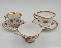 Three vintage teacups and saucers displayed on a white background showing floral designs and gold accents