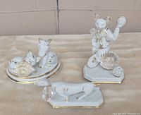 Three white porcelain figurines with gold gilding and Swarovski crystals displayed together on a beige surface.