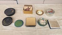 Nine varied coaster sets displayed on a light surface, including horse-themed square coasters, black lacquer Japanese style coasters, woven straw coasters, floral and silver painted coasters in container.