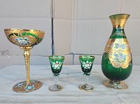 Front view of vintage Murano green and gold vase, cup, and two liquor glasses on cloth-covered surface.